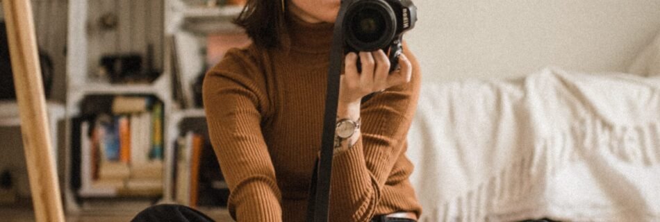 woman sitting on brown wooden floor while holding black DSLR camera in room